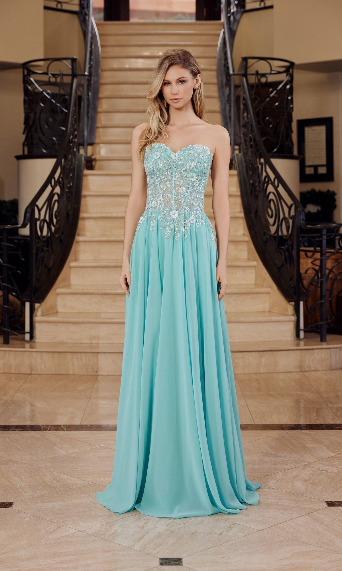 A woman stands indoors by a staircase, wearing the Nox Anabel C1617 Strapless Sweetheart Prom Dress in sage green, featuring a fitted embellished bodice and flowing floor-length skirt.